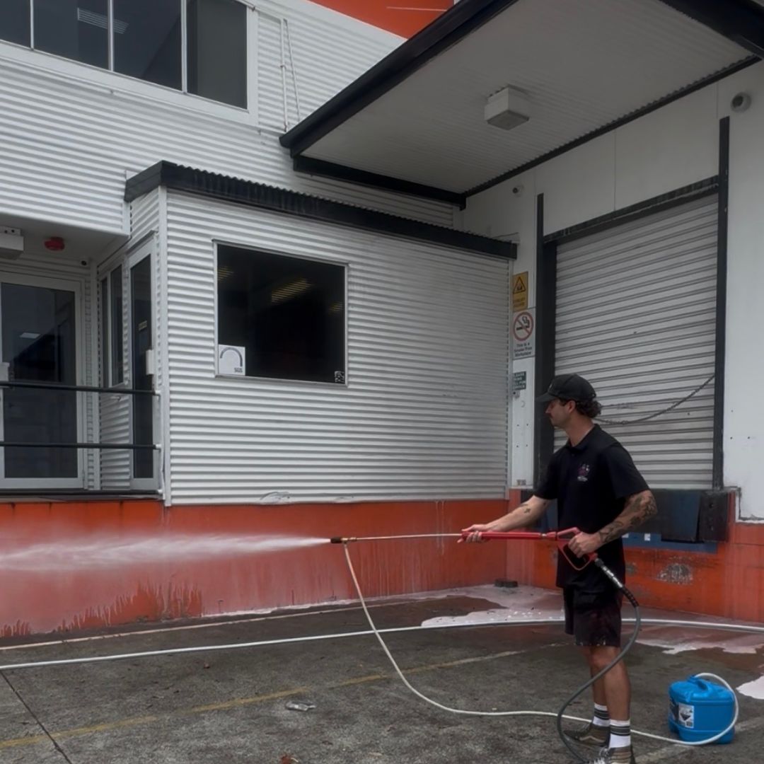 One Call Property Care technician applying chemical treatment via low-pressure wand to the orange-painted lower wall section of the Acit facility — chemical container visible on the ground beside the technician with the corrugated cladding and roller door visible behind