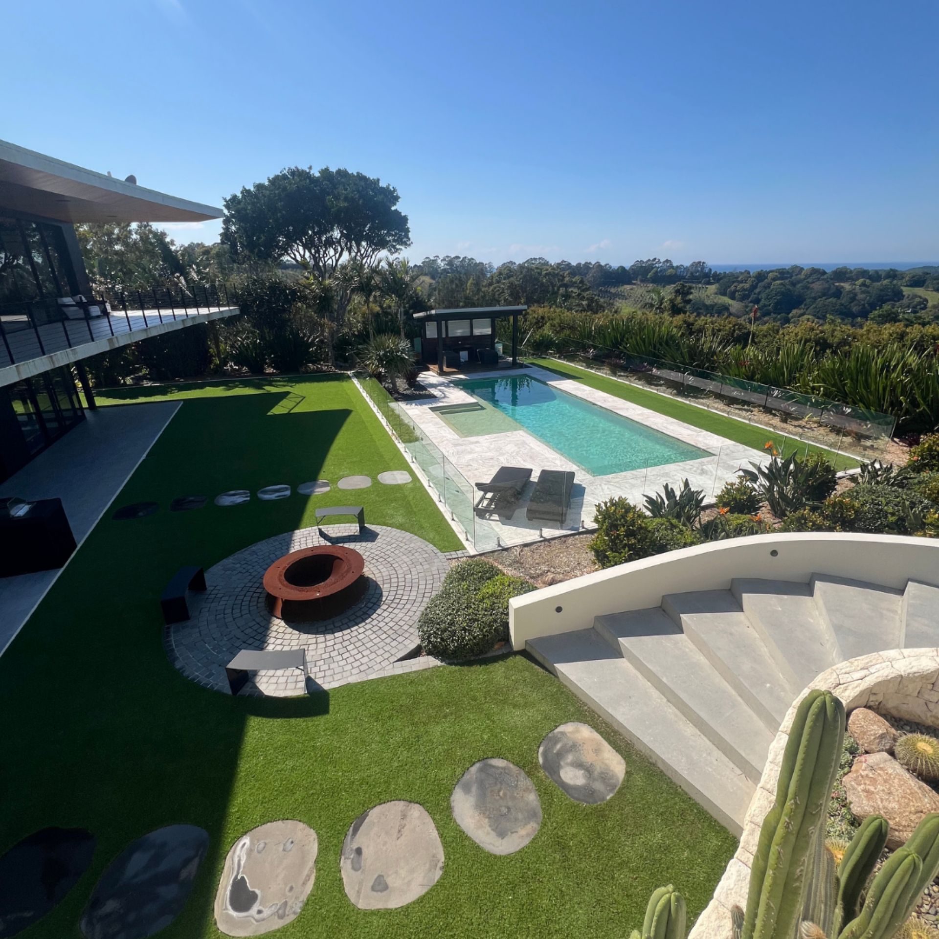 Wide property context shot of the Byron Bay luxury mansion — modern architecture, pool, glass balustrade, fire pit feature, synthetic turf lawn, stepping stones, curved stairs, and ocean view across the Byron hinterland visible in the background