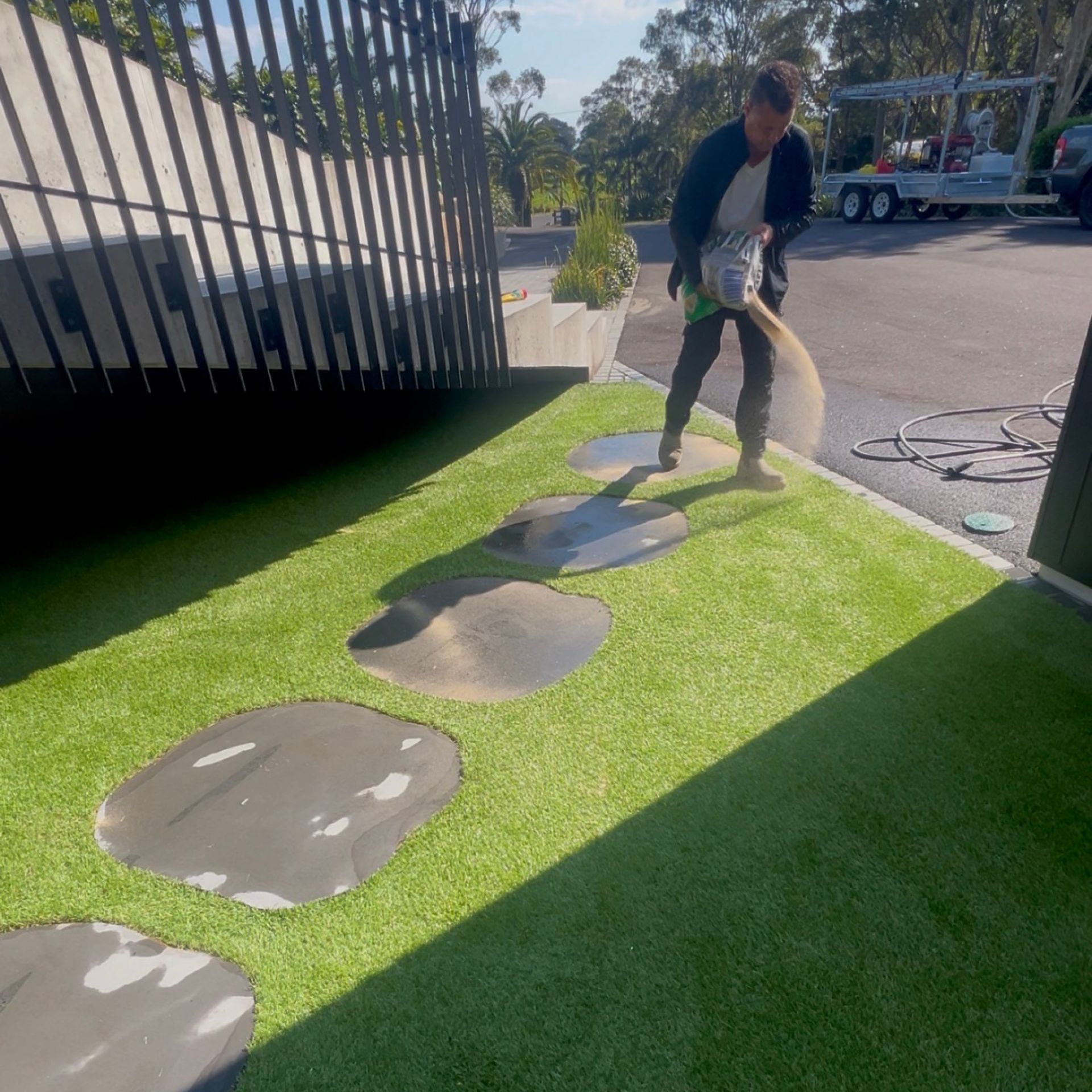 One Call Property Care technician applying fresh infill sand to synthetic turf around stepping stones at a Byron Bay luxury mansion — sand spreading from a bag mid-throw, with the turf and stepping stone feature visible and a work van and trailer in the background