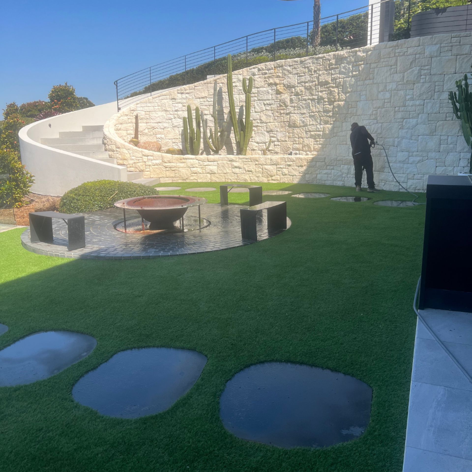 Wide work-in-progress shot at the Byron Bay mansion — One Call technician pressure washing the cream stone retaining wall, with the synthetic turf lawn, fire pit feature, and curved stair detail visible across the landscaped garden area