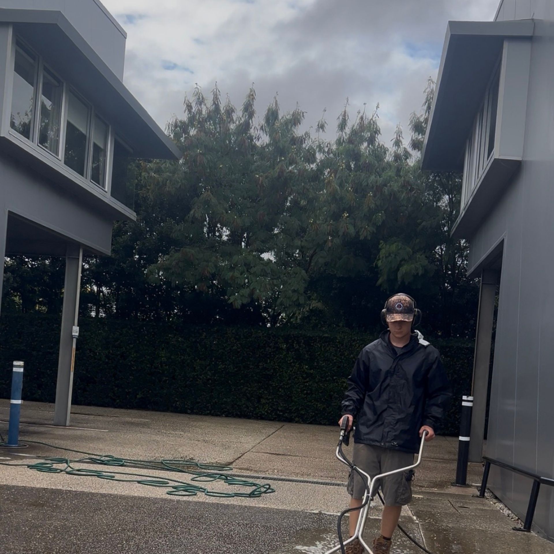 One Call Property Care technician in PPE including ear protection and waterproof jacket operating a surface cleaner attachment on a large pebblecrete commercial forecourt at a physio practice — work in progress with both buildings of the practice visible either side and the wet cleaned section visible behind